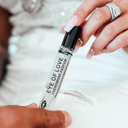 Ein klarer glas-roll-on-flakon mit schwarzem deckel labeled eye of love pheromon parfum wird zwischen zwei händen gehalten