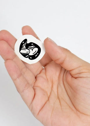 Weißer kreisförmiger aufkleber mit schwarzer illustration von zwei ineinandergreifenden stilisierten fröschen aus der spicy collection temporary tattoos