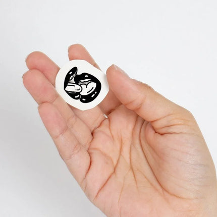 Weißer kreisförmiger aufkleber mit schwarzer illustration von zwei ineinandergreifenden stilisierten fröschen aus der spicy collection temporary tattoos